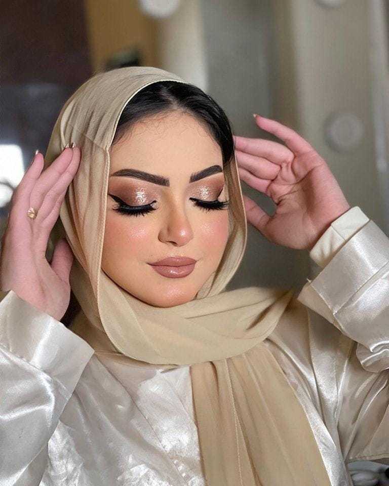 Woman in beige hijab with elegant makeup, shimmering eyeshadow, and glossy lips, eyes closed.